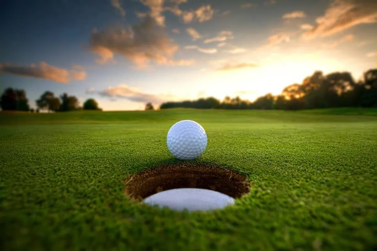 A lively exterior shot of a lush green golf course, with a close-up of a golf ball near the hole. The setting is tranquil, inviting moments of recreation, celebration, and connection with nature at sunset.