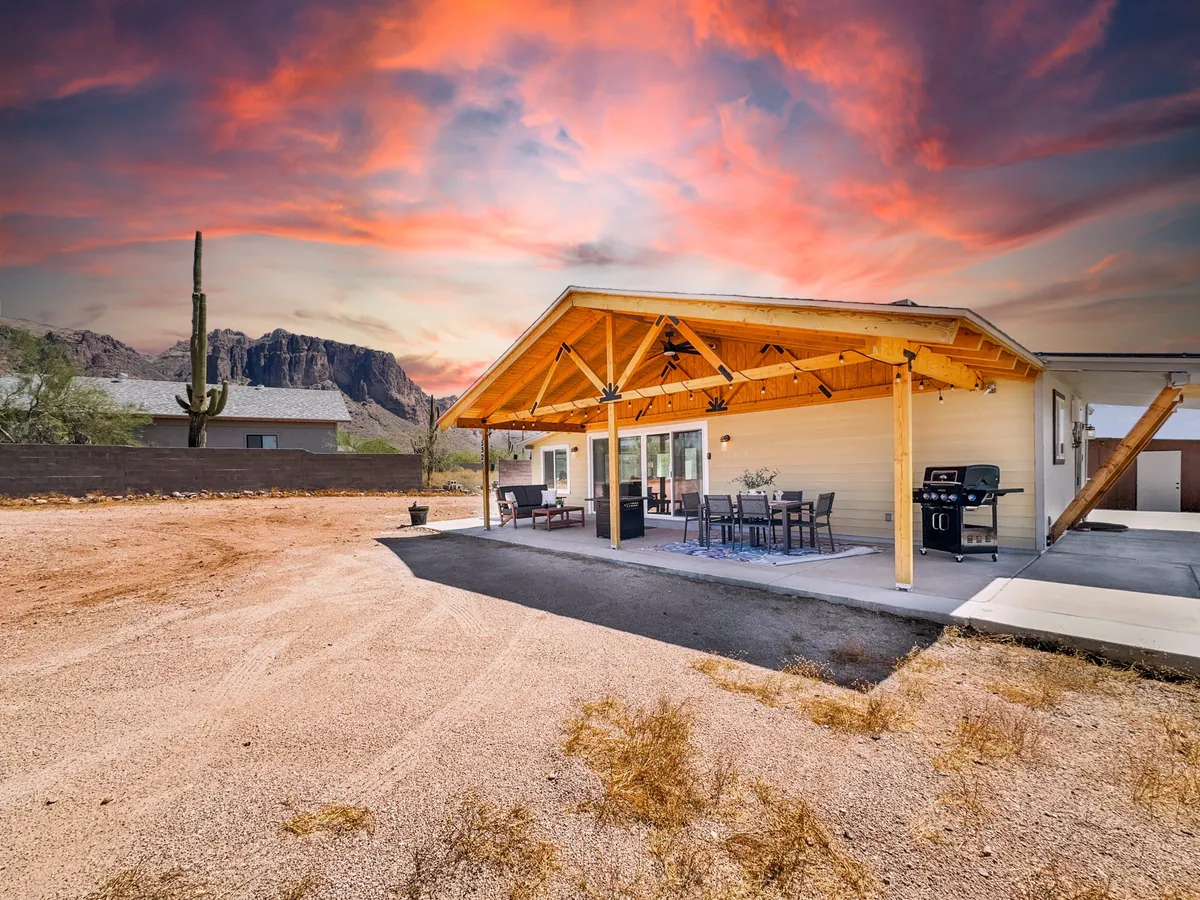 A covered patio featuring a spacious outdoor dining area, grilling station, and comfortable seating, all set against a stunning mountain backdrop. The patio opens directly to the desert landscape, with the Superstition Mountains clearly visible in the distance.