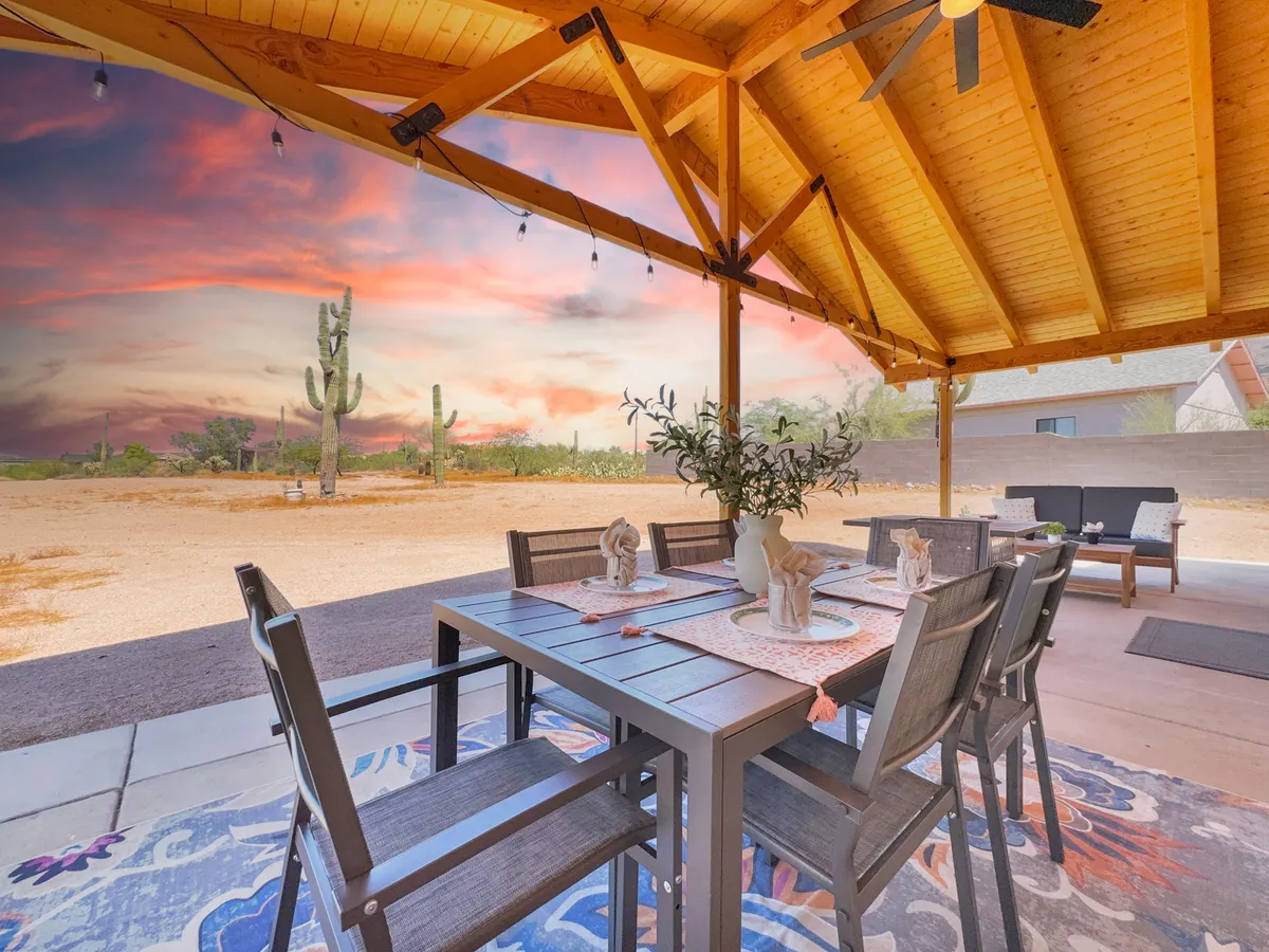 An inviting exterior dining area featuring a rustic wooden table set with a decorative centerpiece and a colorful rug beneath. The scene is surrounded by desert flora, including a prominent cactus in the background, and is bathed in soft evening light, creating an ideal setting for outdoor meals.
