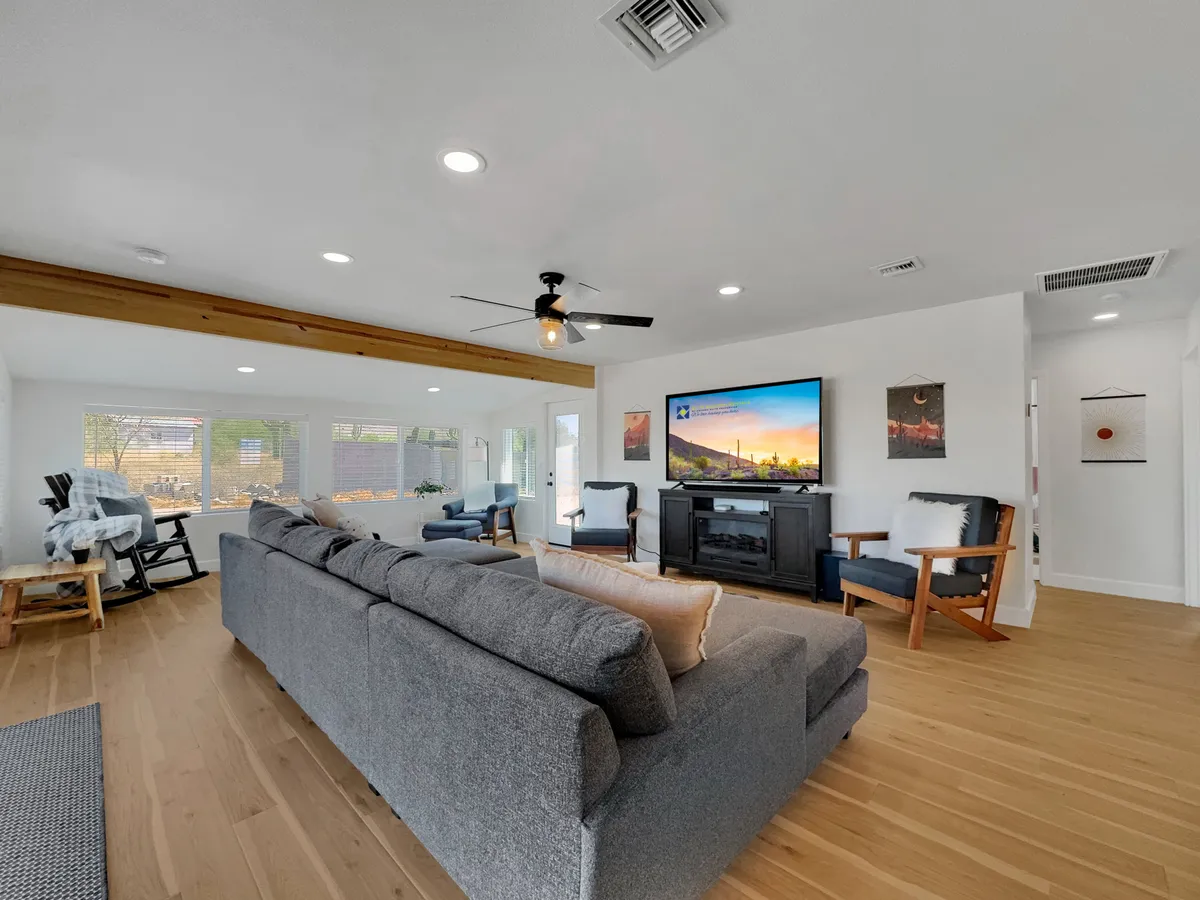 A spacious living room with a large gray sectional sofa, a wall-mounted oversized TV, wooden ceiling beam, and bright windows that let in abundant natural light. The windows reveal hints of the outdoors, emphasizing the blending of interior comfort with the surrounding landscape.