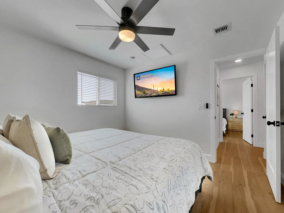 A cozy bedroom with a bed adorned with decorative pillows, a TV mounted on the wall across from the bed, and a window with blinds allowing for adjustable privacy and light. The furnishings and layout emphasize both comfort and practical amenities.