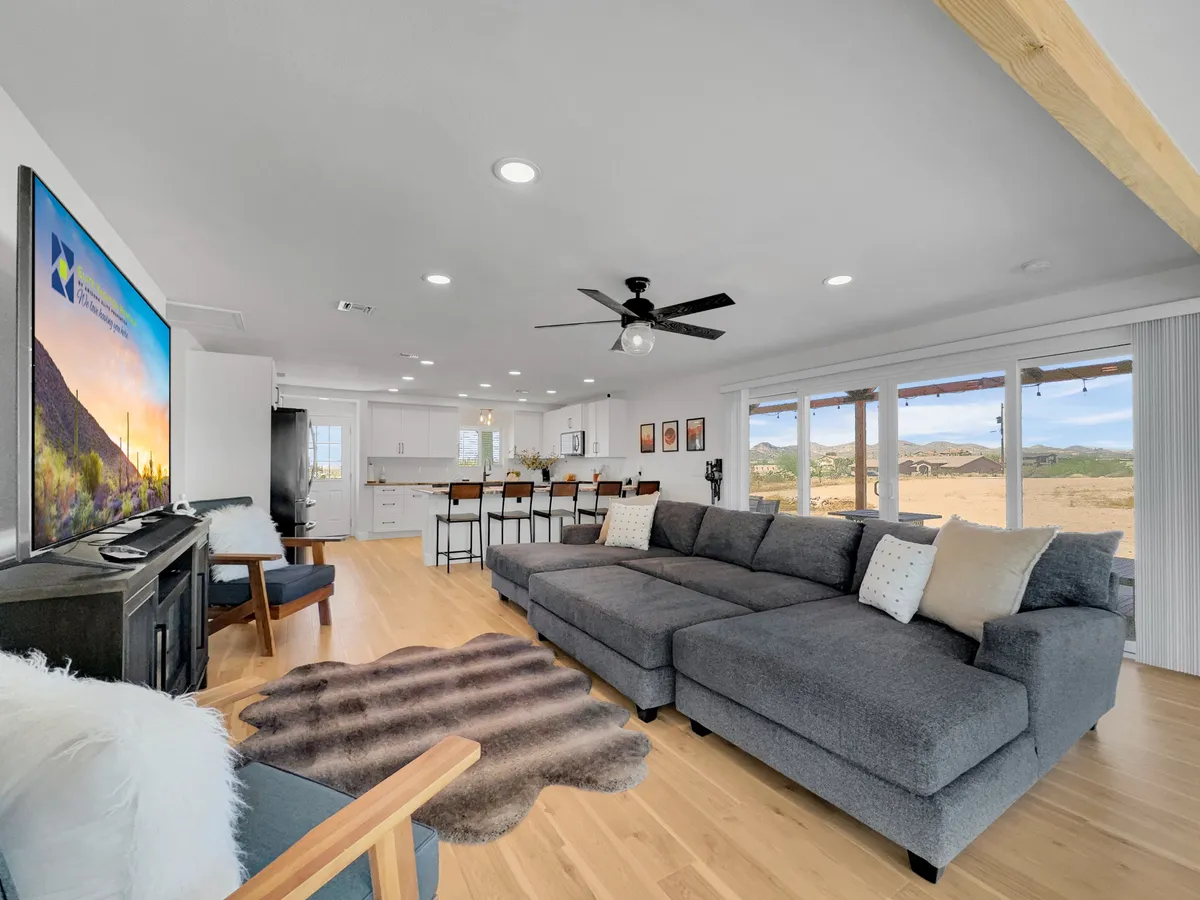 An open-concept living area with a large sectional sofa facing a TV, but also prominently featuring the kitchen island with bar stools. Large windows bathe the space in natural light and offer views of the landscape, enhancing the sense of openness and sociability.