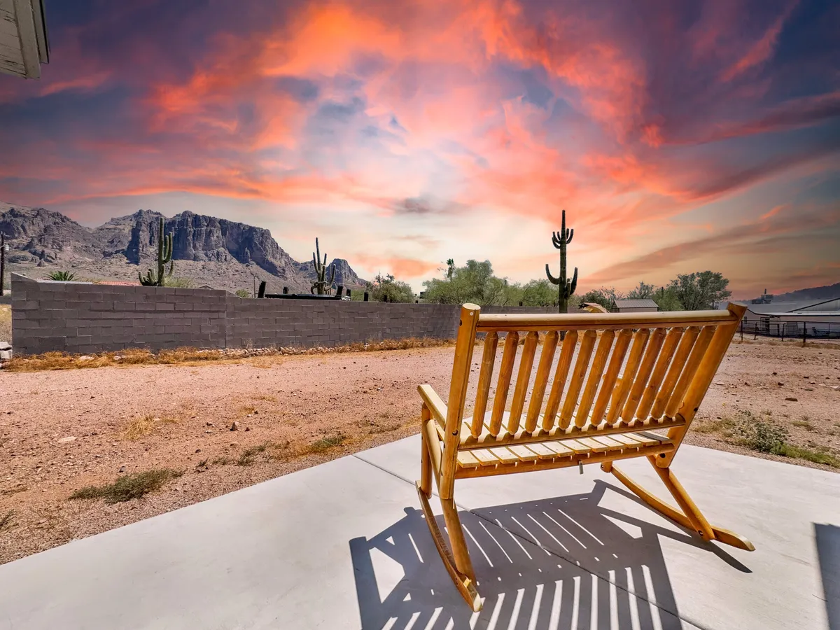 A cozy front porch with a wooden rocking chair facing out toward a dramatic sky and a sweeping mountain range. Cacti dot the landscape, and the porch is sheltered, providing a comfortable spot to soak in the sunset and the natural beauty that surrounds the property.