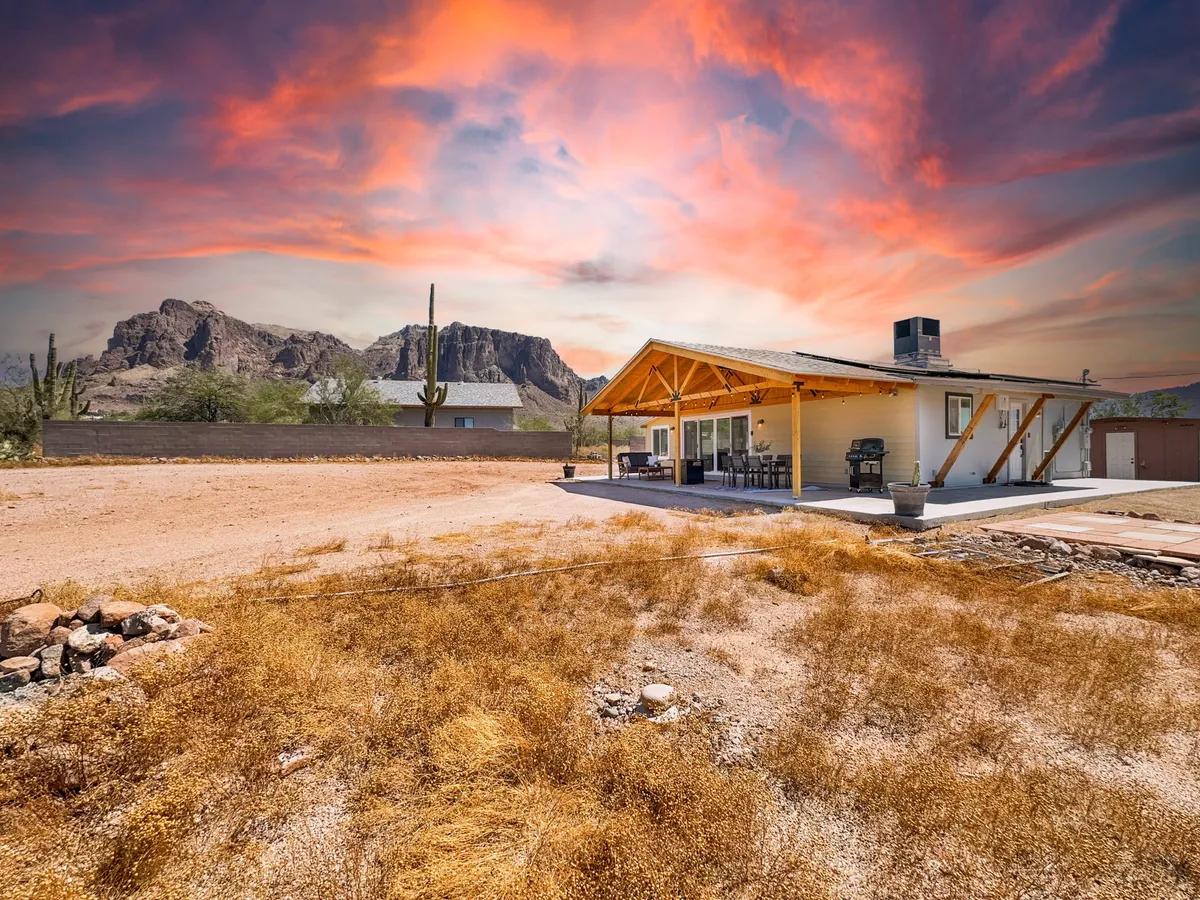 A broad view of the home with a wooden roof, open-air parking area, and a mountain backdrop. The surrounding open space and dry grass evoke the rural Arizona setting, emphasizing adventure and tranquility.