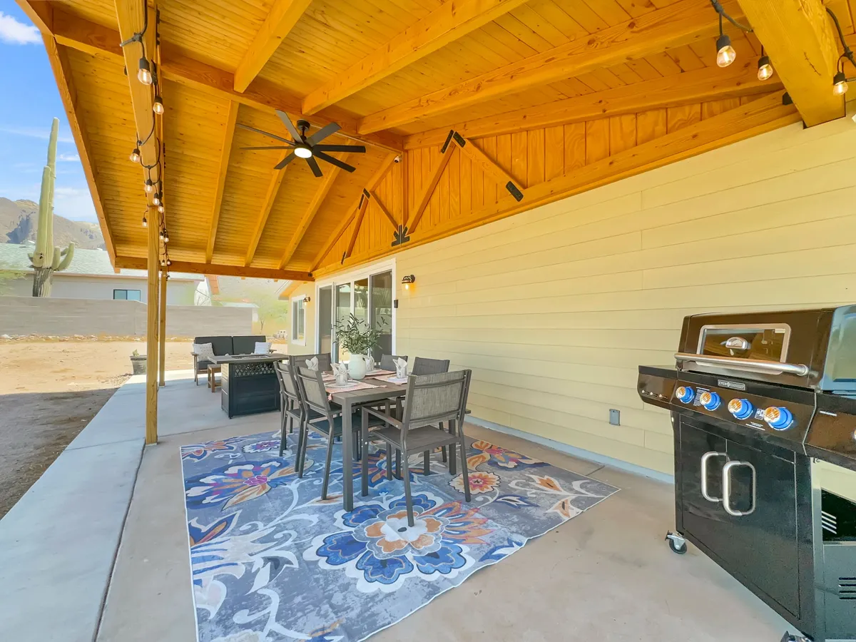 An inviting exterior patio setup featuring a dining table, grill, decorative rug, and a wooden ceiling with a cactus in the background. The scene exudes a casual, welcoming vibe ideal for post-event relaxation, outdoor meals, or gatherings with friends and family.